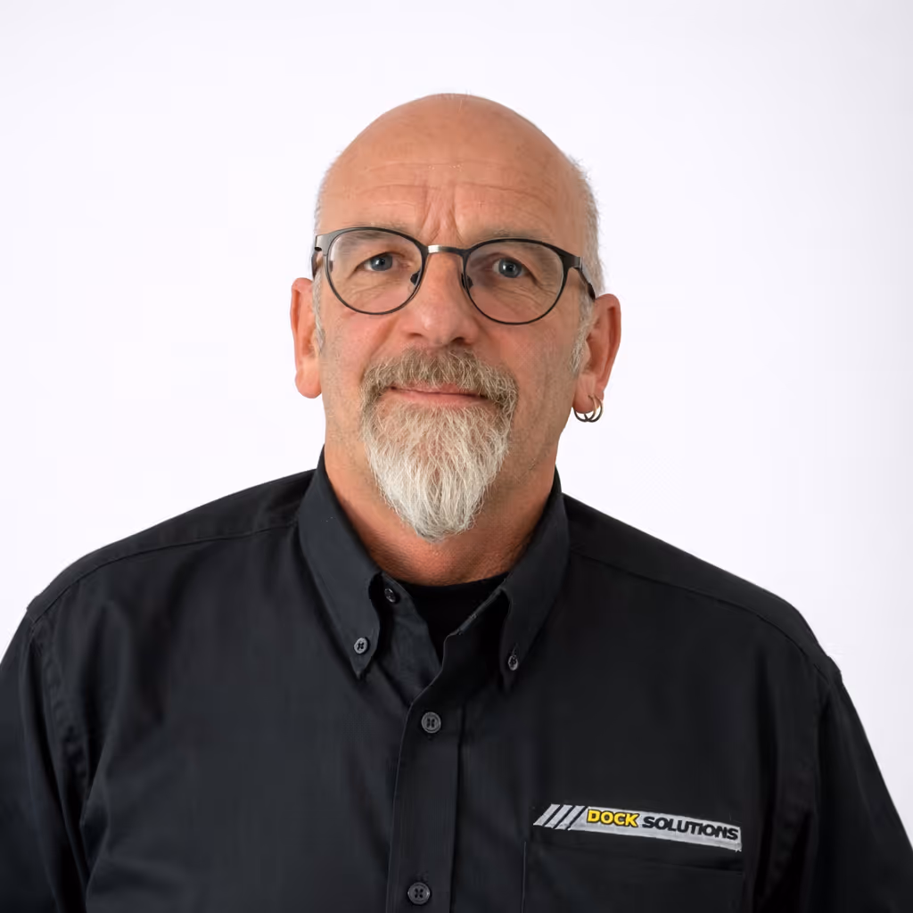 Portrait of a middle-aged bald man with glasses, a white goatee beard, and a hoop earring wearing a black shirt with a Dock Solutions logo on the chest pocket.