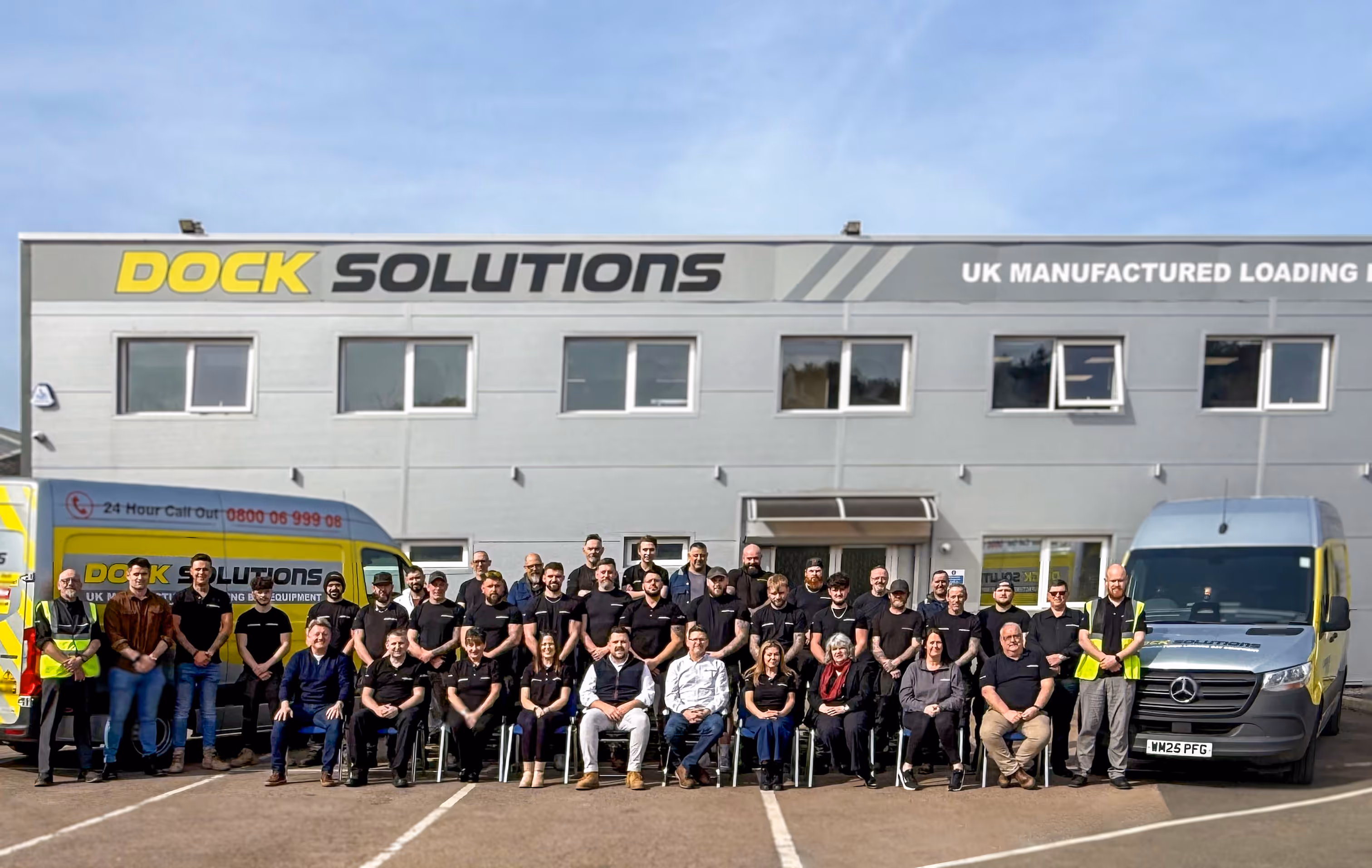 Group photo of Dock Solutions team standing and sitting outside a building with Dock Solutions signage and two company vans on either side.