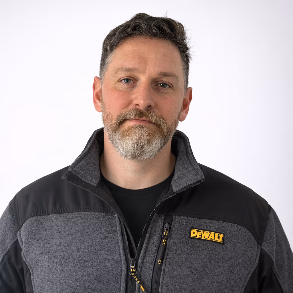 Man with short gray and brown hair and a beard wearing a gray and black jacket with a yellow DeWalt logo on the chest, standing against a plain white background.