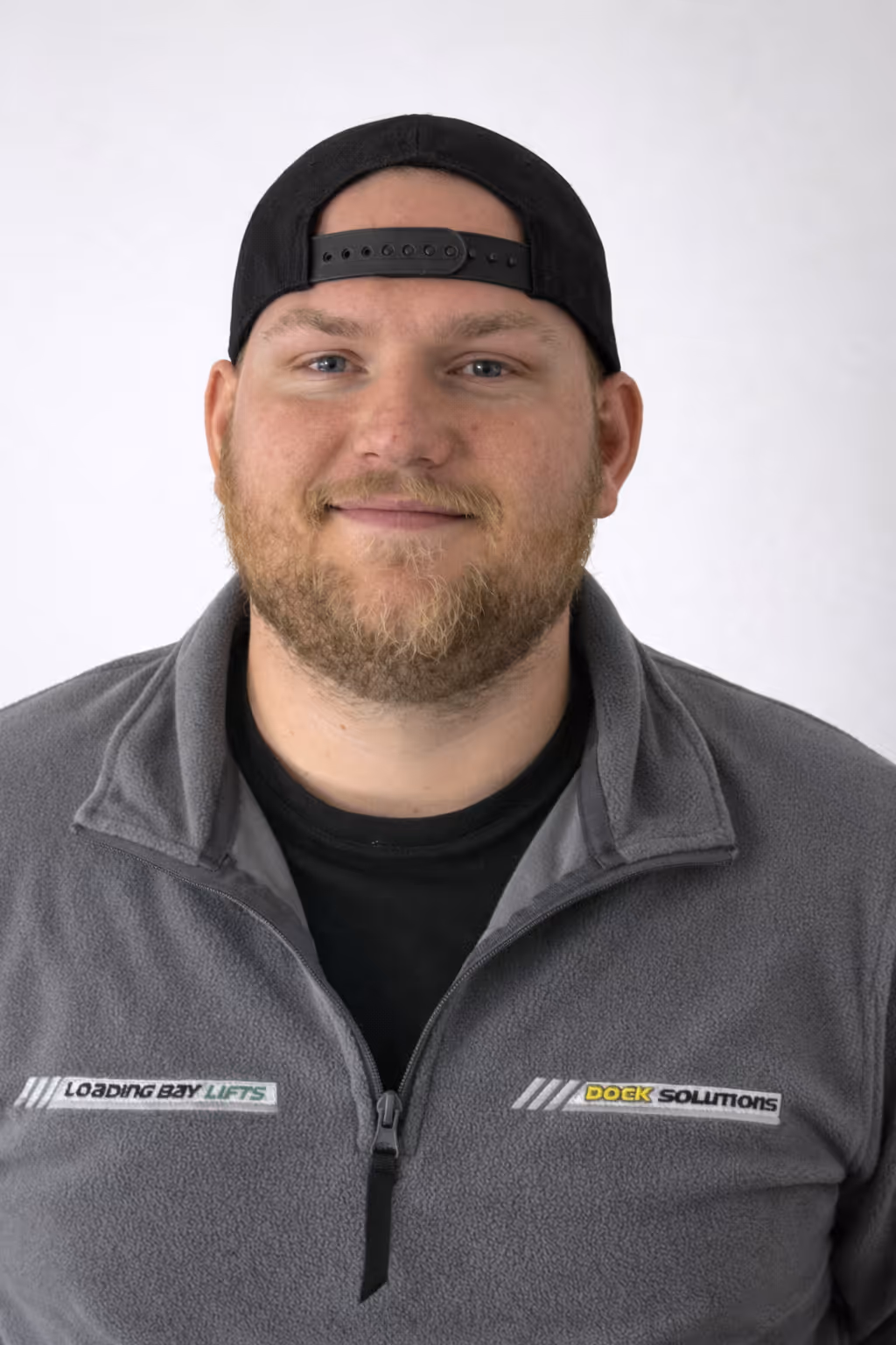 Man with a beard wearing a black backward baseball cap and a gray jacket with logos reading 'Loading Bay Lifts' and 'Dock Solutions' on a white background.