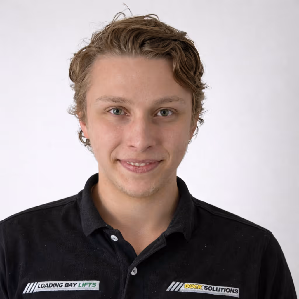 Young man with light brown wavy hair wearing a black collared shirt with logos 'Loading Bay Lifts' and 'Dock Solutions' on the chest, smiling against a plain light background.