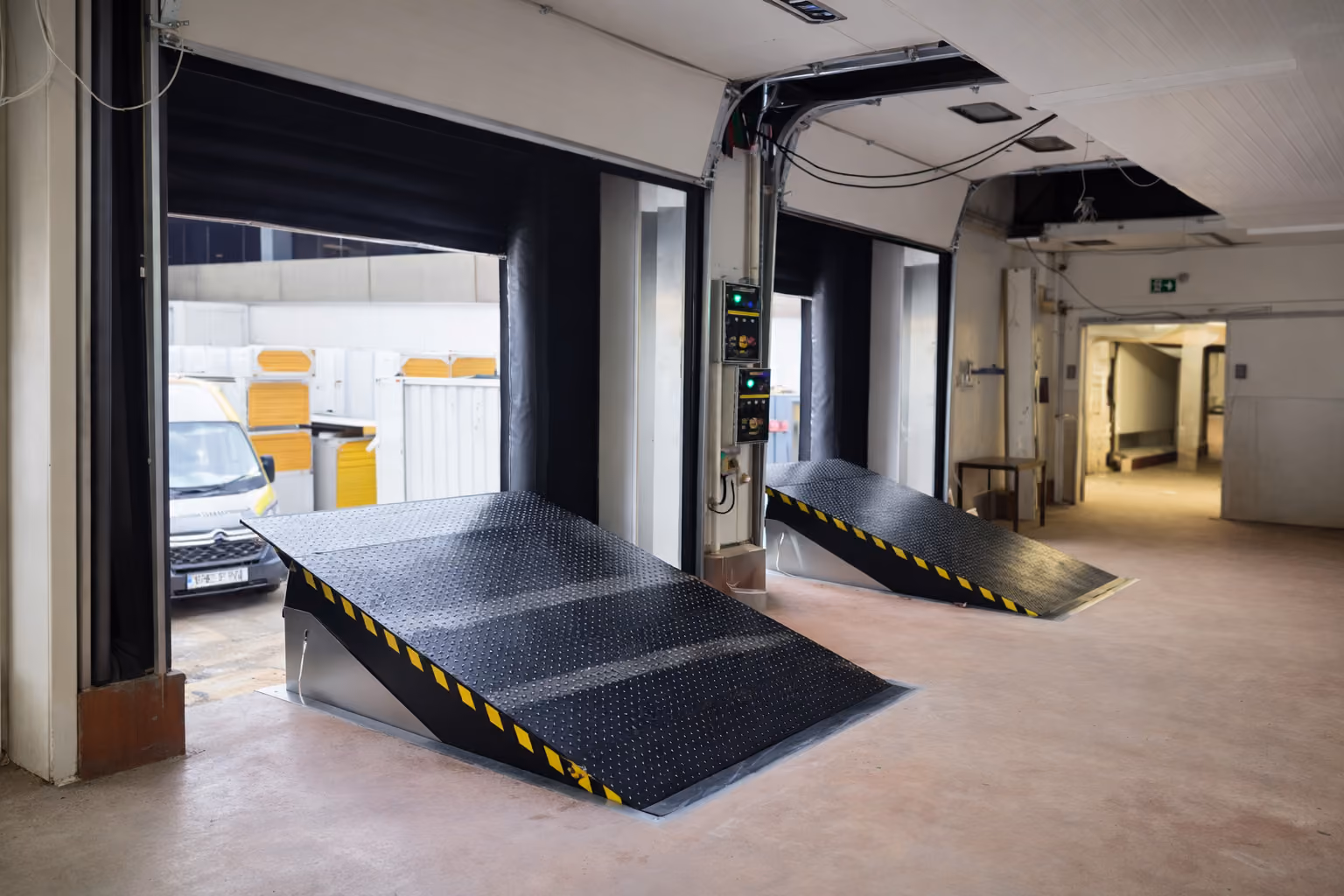 Two black, textured metal loading dock ramps with yellow and black hazard stripes inside a loading bay with open garage doors, showing a white van and shipping containers outside.
