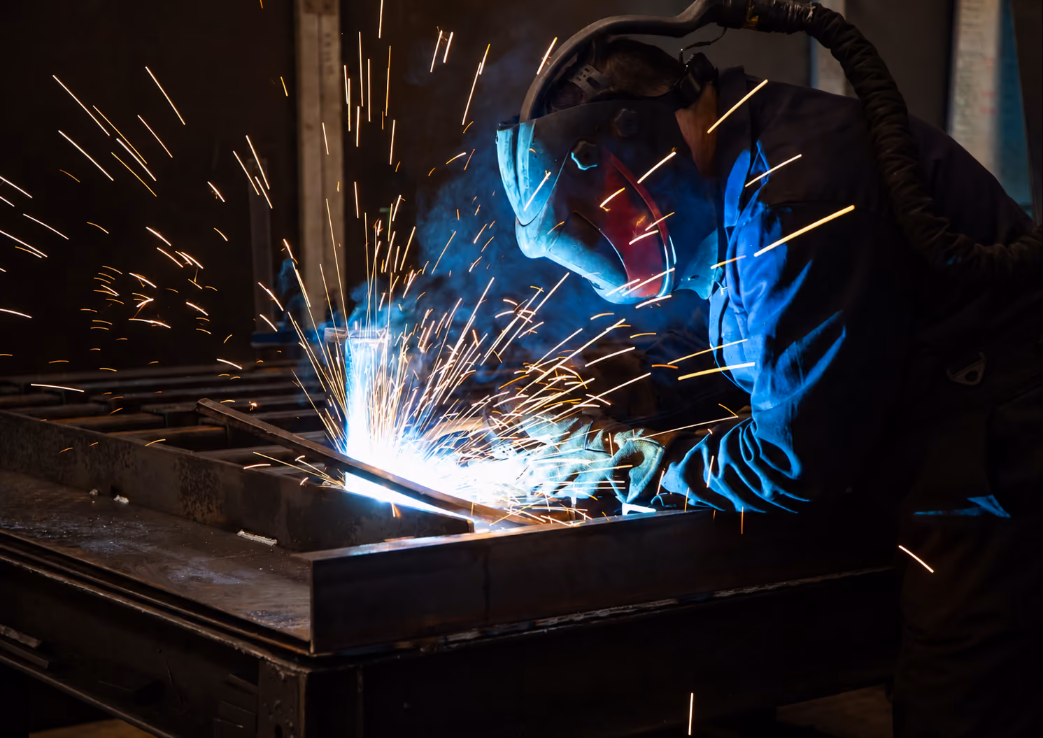 Person wearing protective welding helmet and gloves, working on metal with sparks flying in a dark workshop.