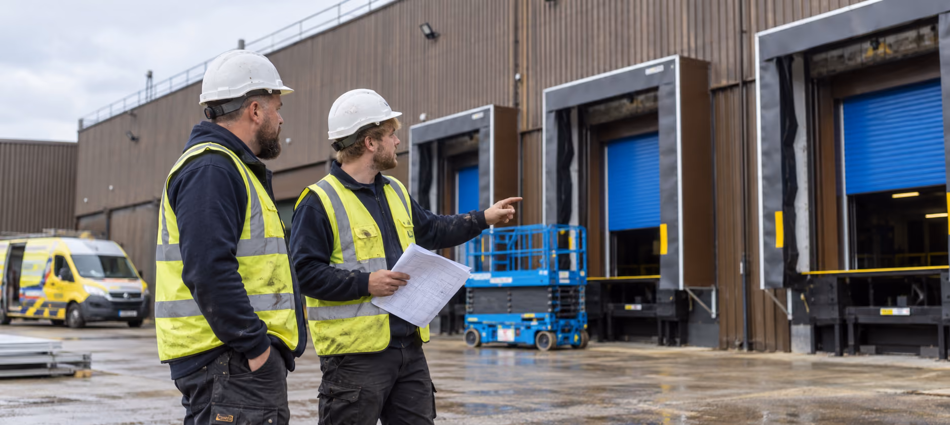 Two construction workers wearing white helmets and yellow safety vests at an industrial site; one worker holds papers and points towards three large loading dock doors with blue shutters, and a yellow van and blue scissor lift are visible in the background.