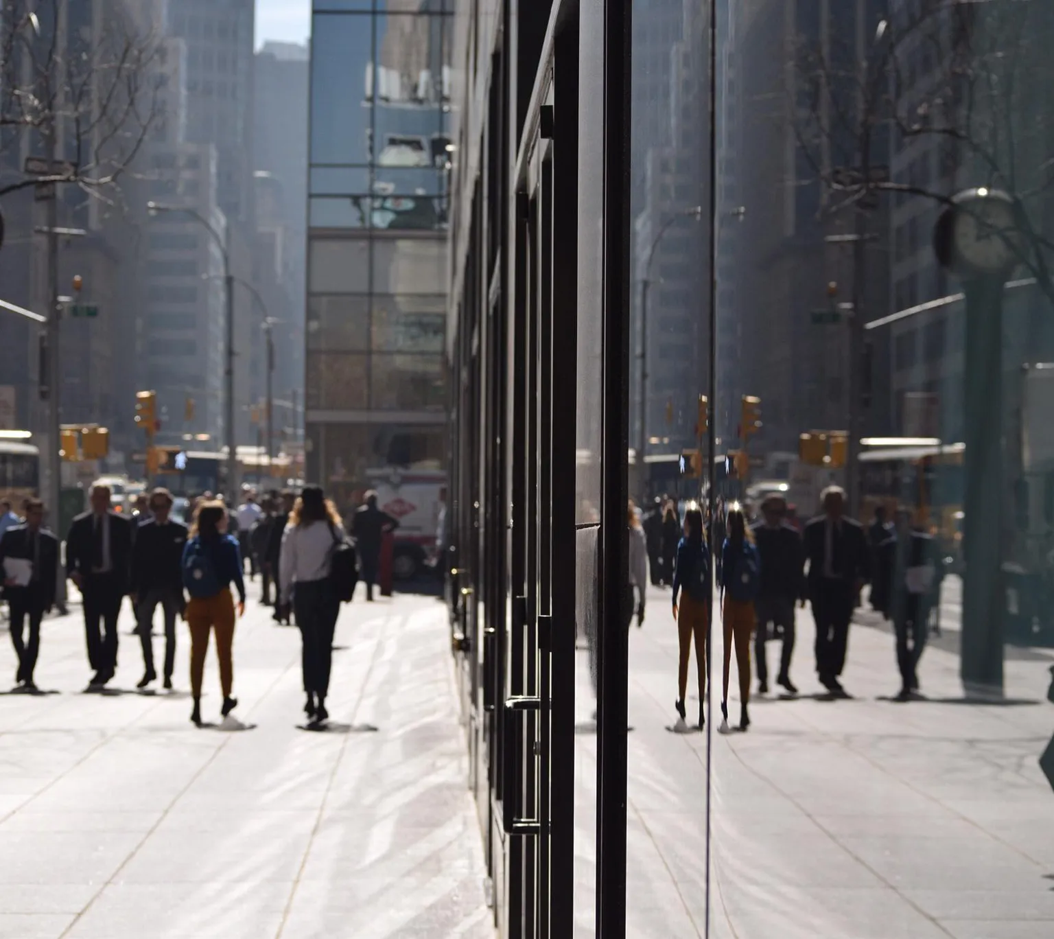 City sidewalk busy with people walking beside a reflective glass building under sunlight.
