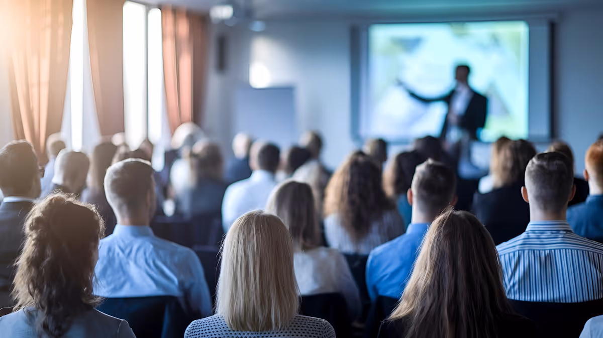 Audience seated facing a presenter giving a lecture or presentation in a conference room.