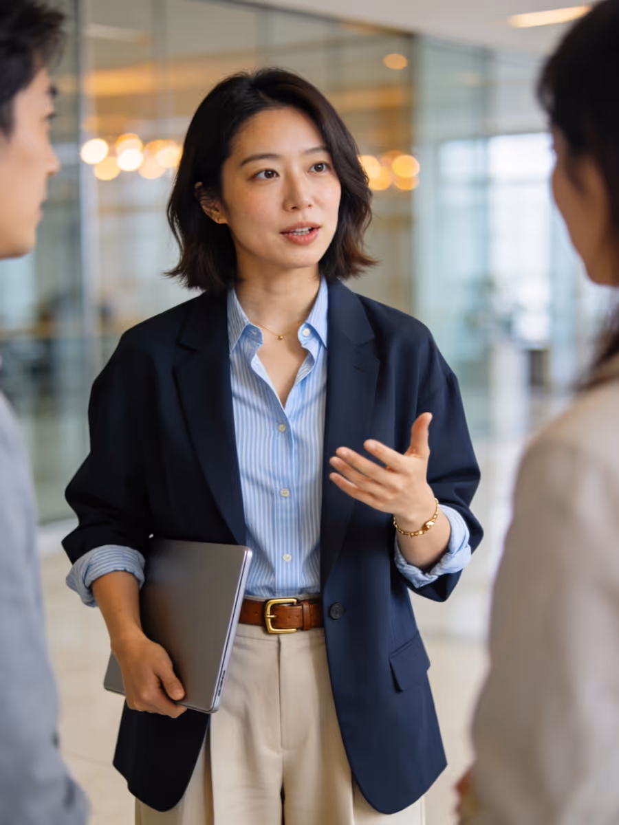 Businesswoman in a navy blazer holding a laptop and speaking to colleagues in an office setting.