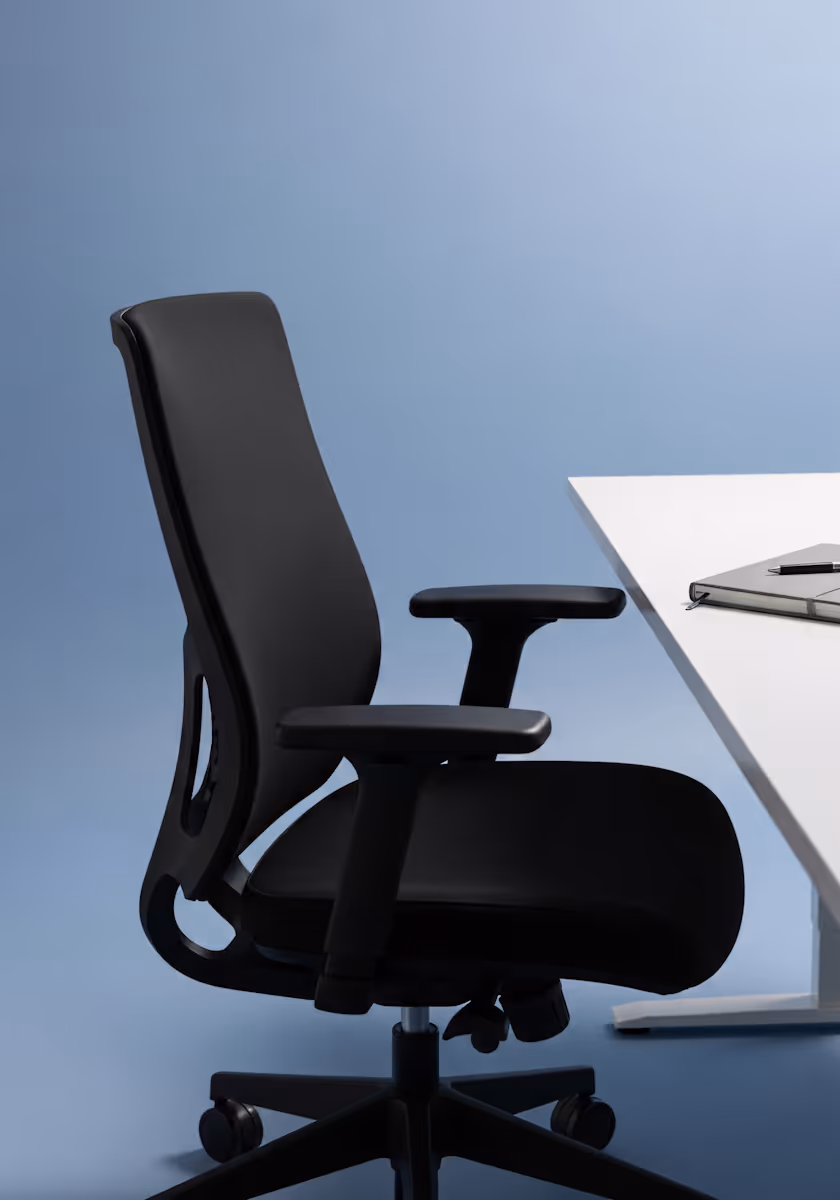 Black ergonomic office chair with adjustable armrests positioned next to a white desk with a closed notebook and pen.