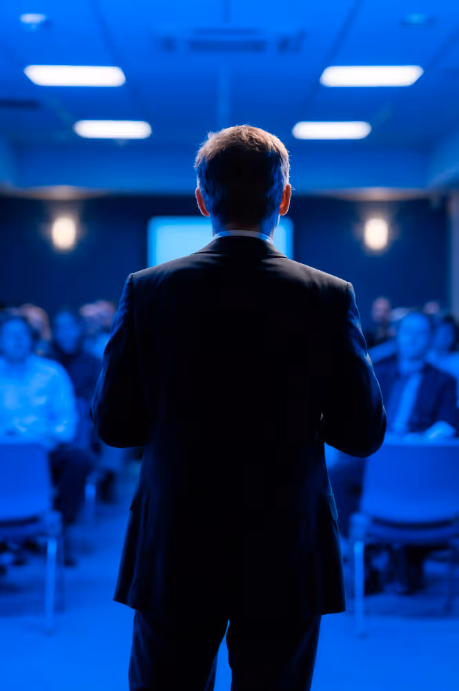 Man in a suit seen from behind giving a presentation to an audience in a dimly lit conference room with blue lighting.