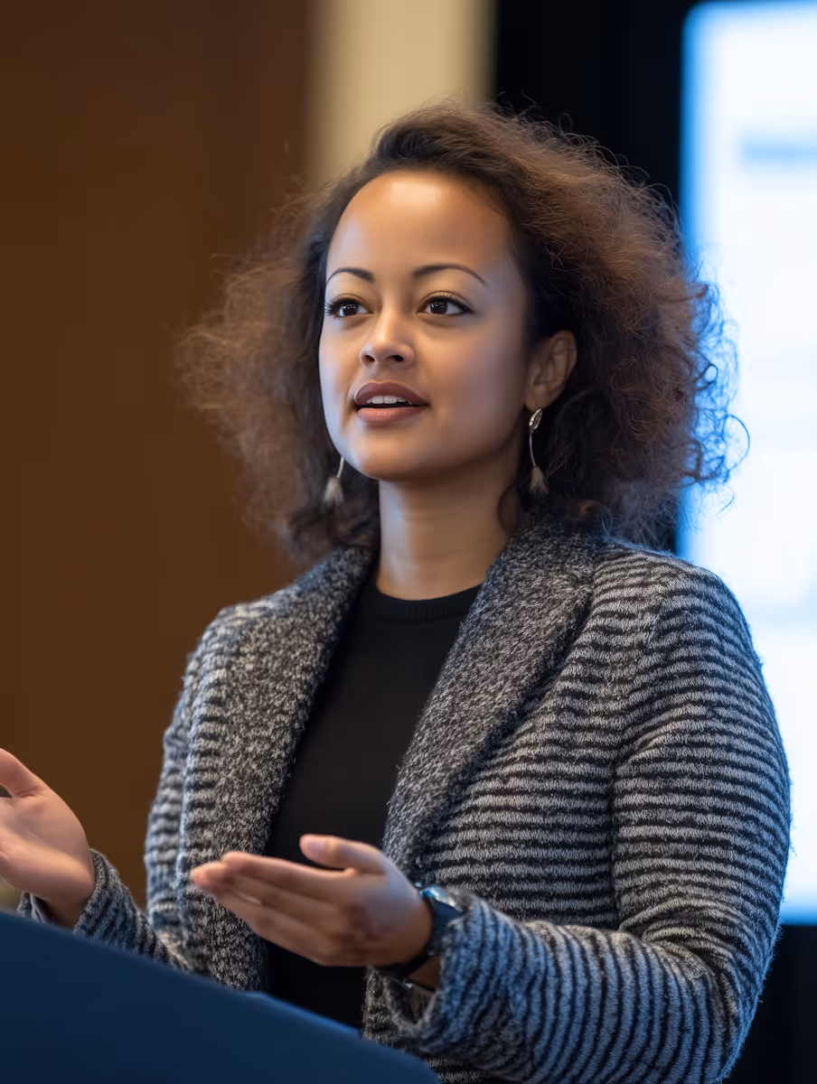 Woman with curly hair wearing a gray striped blazer speaking and gesturing with her hands.
