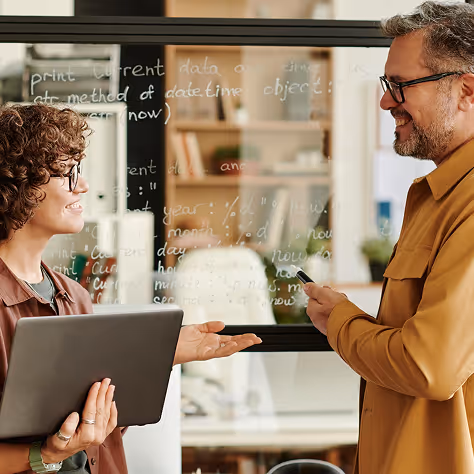 Two colleagues wearing glasses discussing in front of a glass wall with handwritten code.