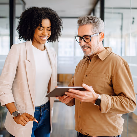 Two colleagues smiling and looking at a tablet in a bright office space.