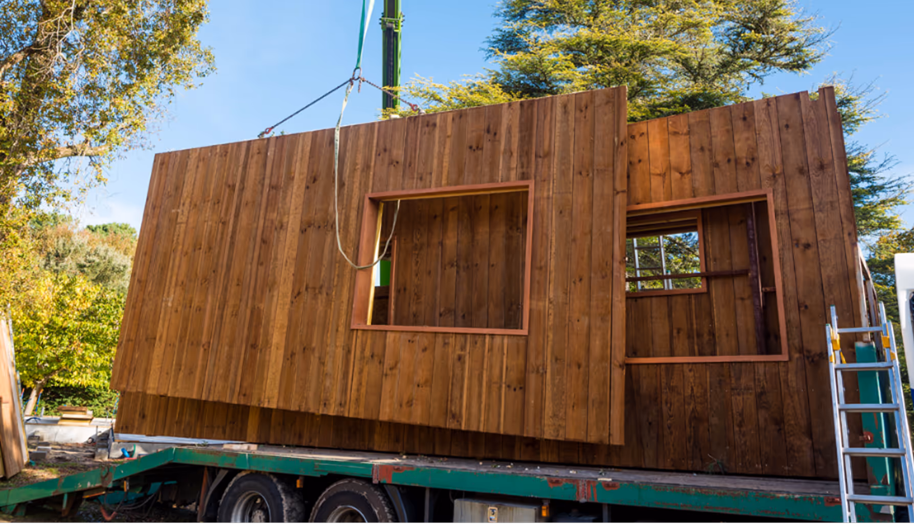 Zwei große vorgefertigte Holzwandabschnitte mit Fensteröffnungen werden von einem Kran auf einen grünen Transportanhänger gehoben.