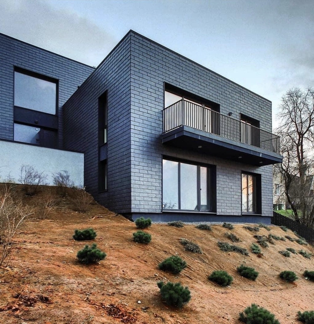 Modern two-story house with dark brick exterior on a slope with small shrubs and trees around.