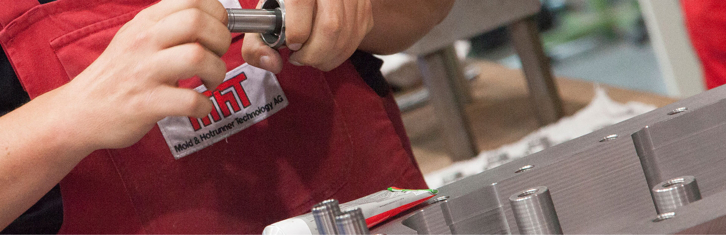 Hände eines MHT-Mitarbeiters montieren ein metallisches Bauteil an einem Spritzguss-Werkzeug. Die Person trägt eine rote Latzhose mit MHT-Logo auf der Brusttasche.