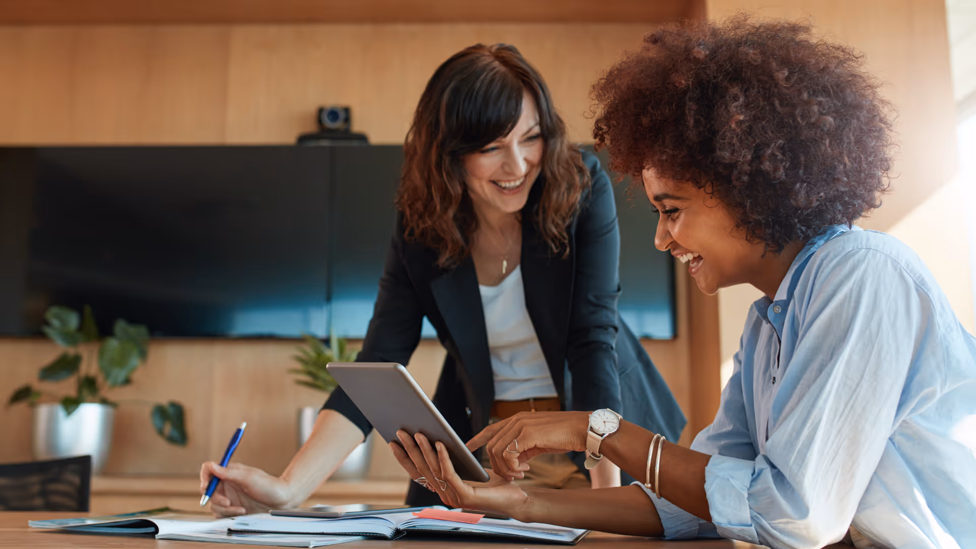 Two women in a modern office smiling and looking at a tablet while discussing work.