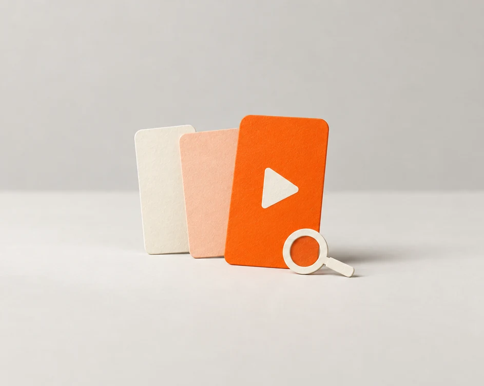 Three paper short-video screen cards in cream, peach and orange, the orange one with a white play button, next to a small cream magnifying glass