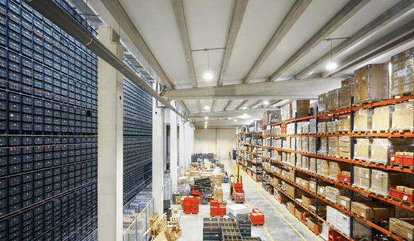 Interior of a large warehouse with shelves stocked with boxes and crates, and some pallets on the floor.