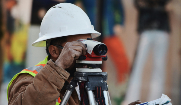 Construction worker in a white hard hat and reflective safety vest using a surveying instrument on a tripod.