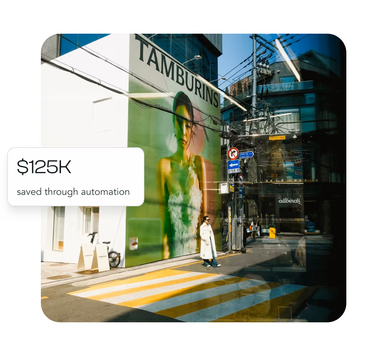 Person in a white coat standing by a crosswalk near a building with a large mural of a woman and 'TAMBURINS' sign.