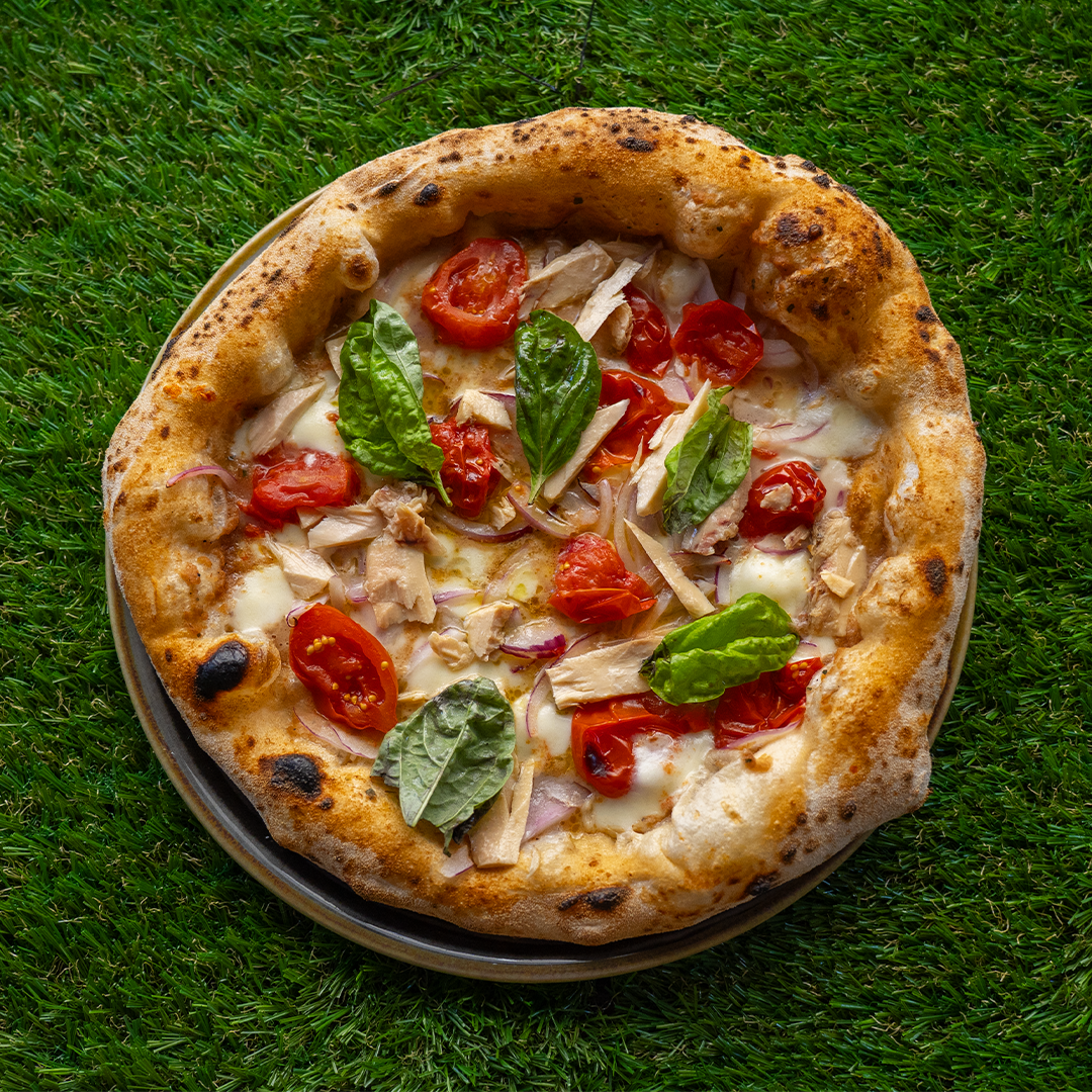 Pizza con tonno, cipolla rossa, pomodorini e foglie di basilico su uno sfondo di erba verde.