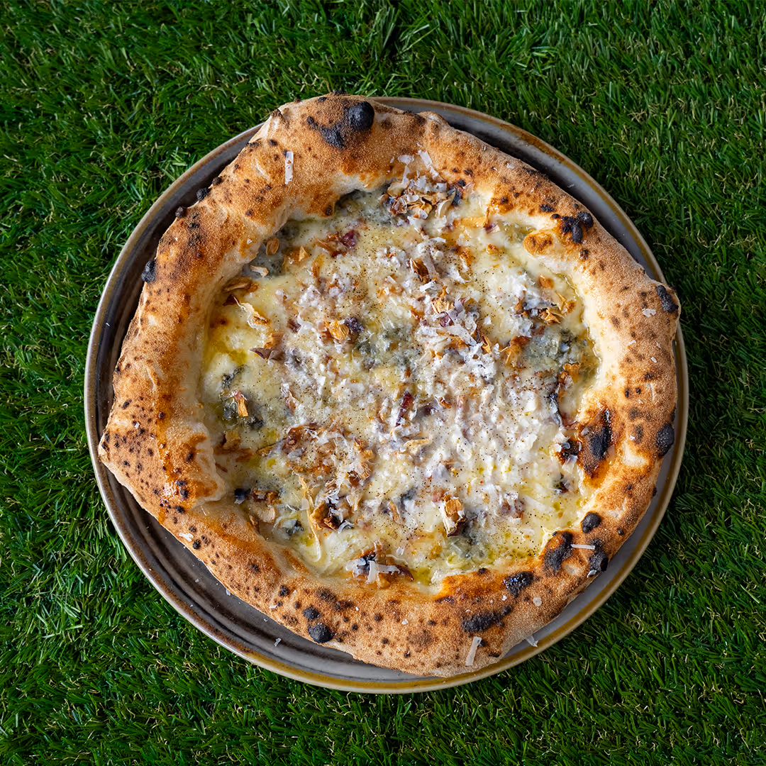 Pizza con crosta dorata e leggermente bruciata, coperta di formaggi fusi e scaglie di formaggio su un piatto grigio, su uno sfondo di erba verde.
