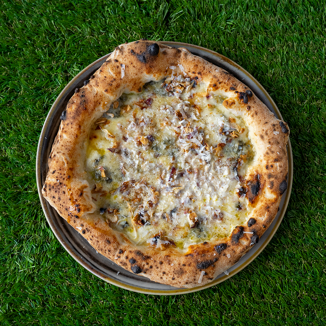 Pizza con crosta dorata e leggermente bruciata, coperta di formaggi fusi e scaglie di formaggio su un piatto grigio, su uno sfondo di erba verde.