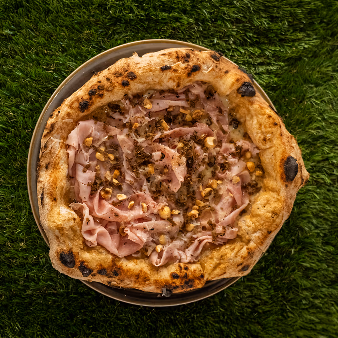 Pizza con prosciutto, nocciole tostate e formaggio su un piatto, poggiata su erba verde.