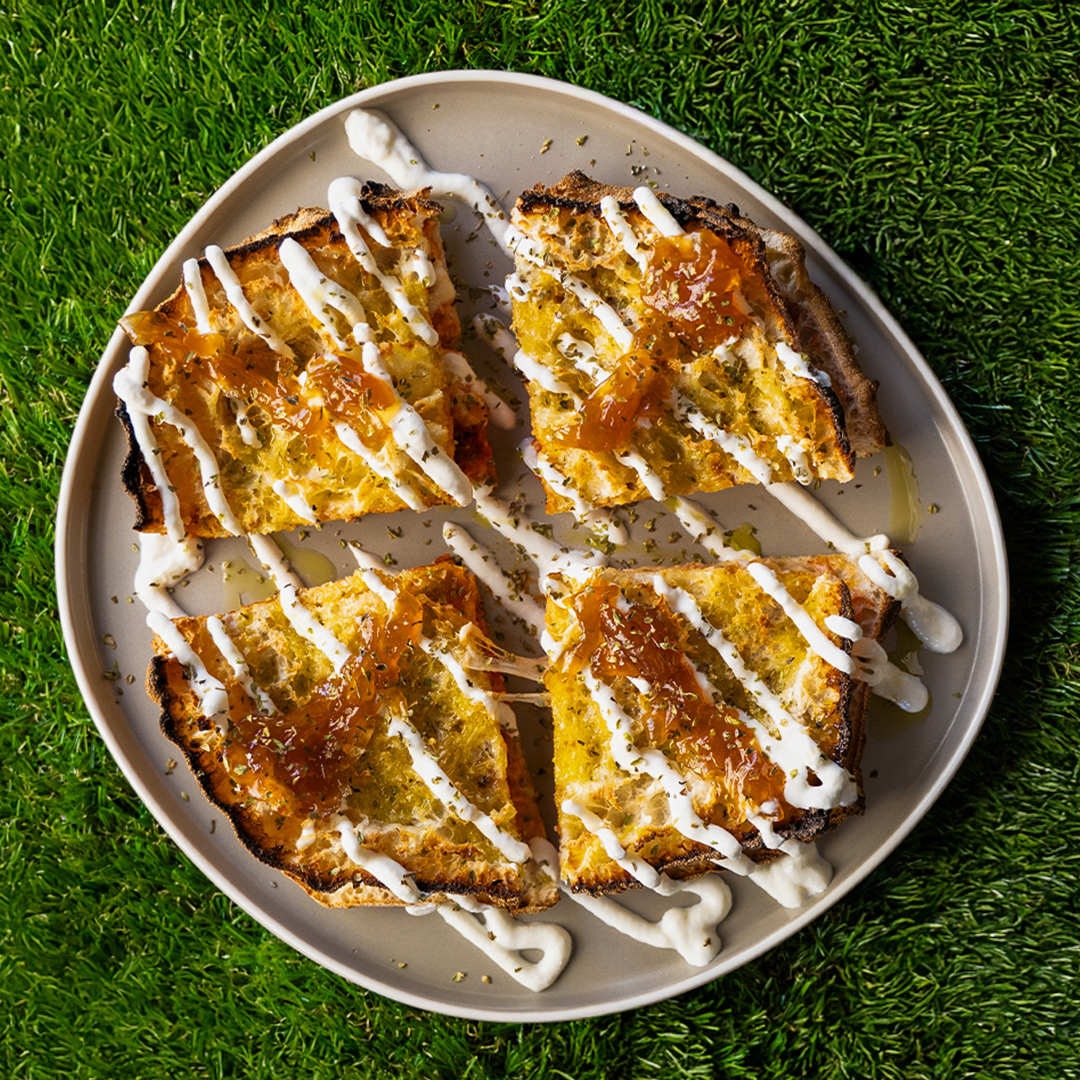 Piatto con quattro fette di pane tostato condita con salsa bianca a strisce e marmellata arancione su sfondo di erba verde.