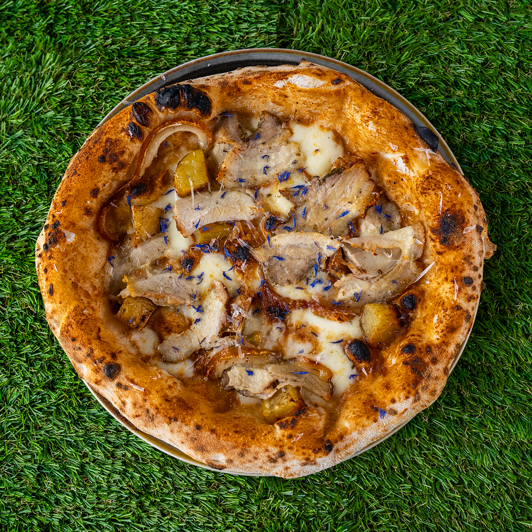 Pizza con fette di porchetta, formaggio fuso, patate e cipolle su uno sfondo di prato verde.