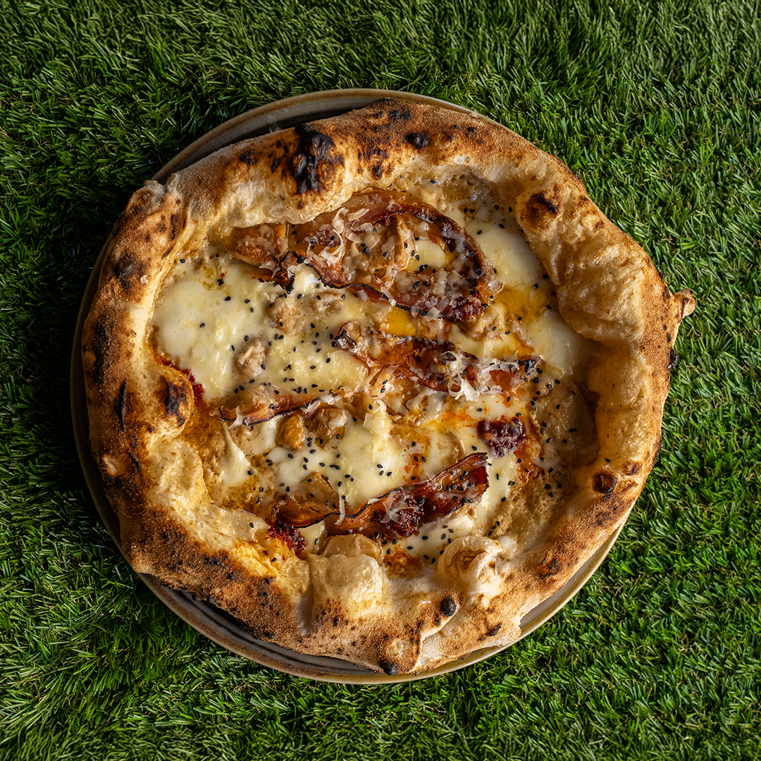 Pizza con crosta spessa e leggermente bruciata, con formaggio fuso, noci, pezzi di pancetta e semi neri su un piatto posato su erba verde.