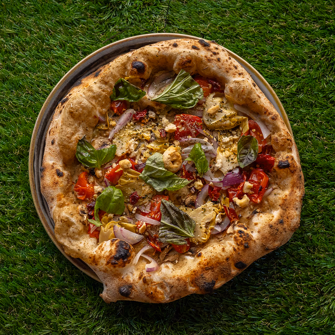 Pizza con crosta spessa e croccante con foglie di basilico, cipolla rossa, pomodori, carciofi e altre verdure su un piatto posto su prato verde.