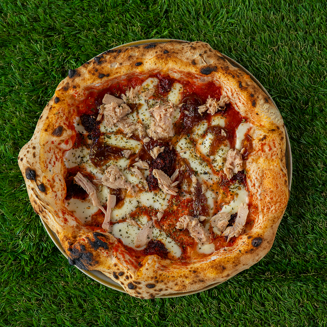 Pizza napoletana con pomodoro, mozzarella, tonno e cipolle caramellate su un piatto sopra erba verde.