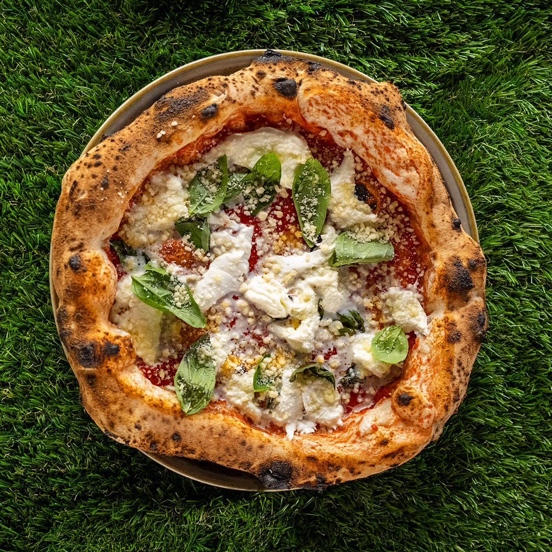 Pizza margherita con crosta dorata e bruciata, mozzarella, salsa di pomodoro e foglie di basilico su piatto, visto dall'alto su sfondo di erba.