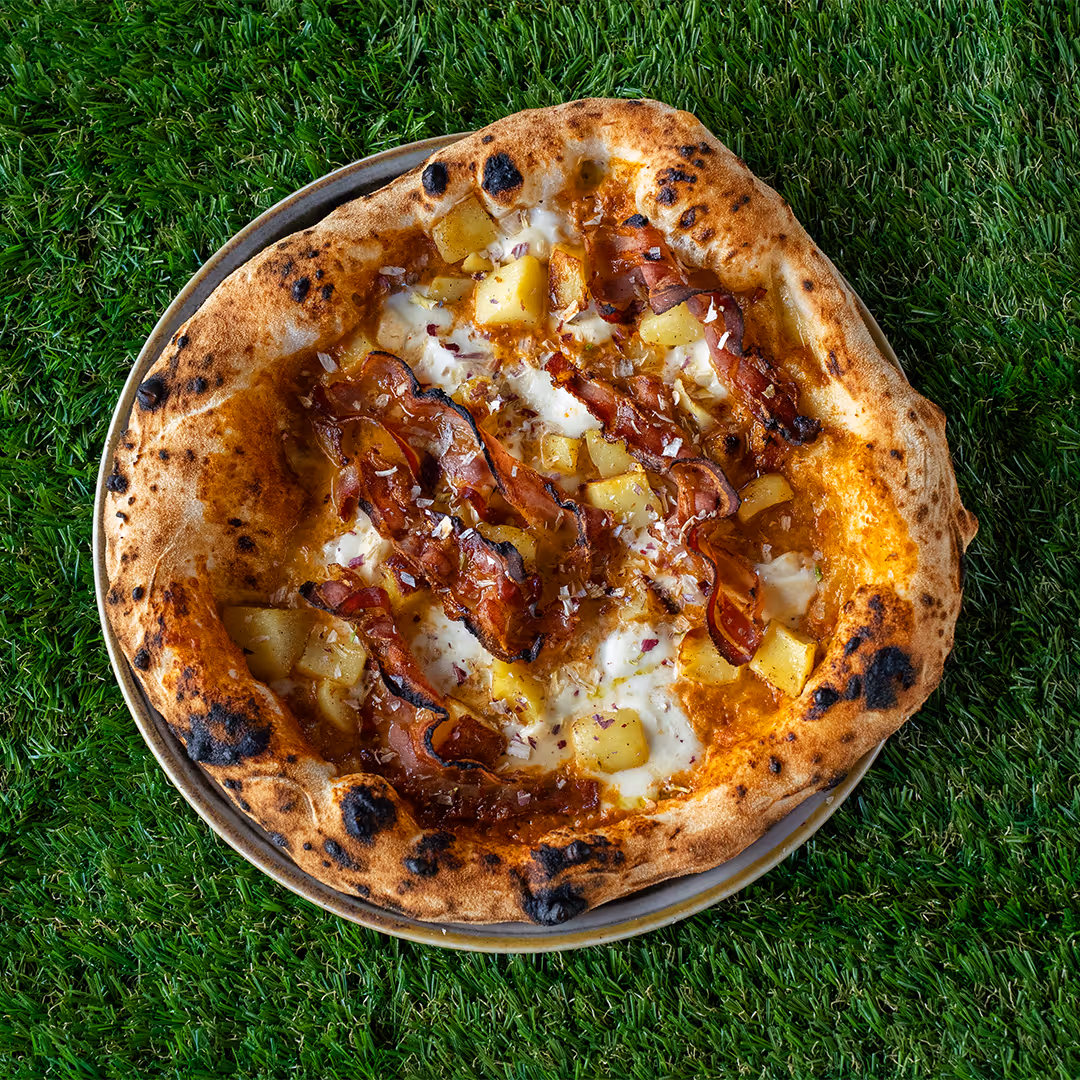 Pizza con bordi dorati e leggermente bruciacchiati con patate a cubetti, mozzarella fusa e fette di pancetta croccante, servita su un piatto su sfondo di erba verde.