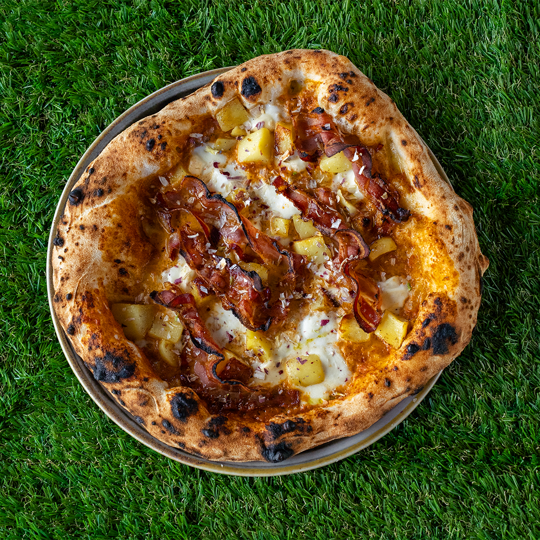 Pizza con crosta dorata, formaggio fuso, strisce di pancetta croccante e cubetti di patate su un piatto, posta su erba verde.
