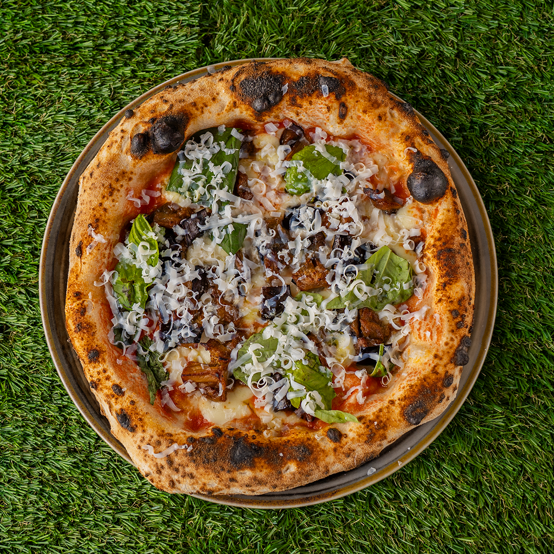 Pizza con crosta leggermente bruciata, condita con salsa di pomodoro, formaggio grattugiato, foglie di basilico e funghi su un piatto, su sfondo di erba artificiale.