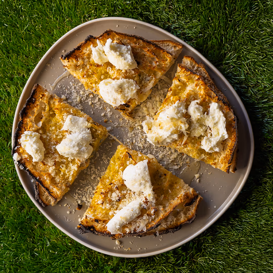 Fette di pane tostato con formaggio spalmabile e scaglie di formaggio su un piatto grigio, poggiato su erba verde.