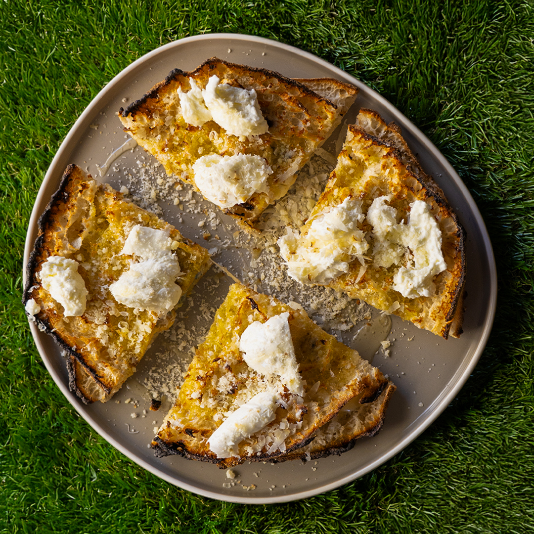 Fette di pane tostato con formaggio spalmabile e scaglie di formaggio su un piatto grigio, poggiato su erba verde.