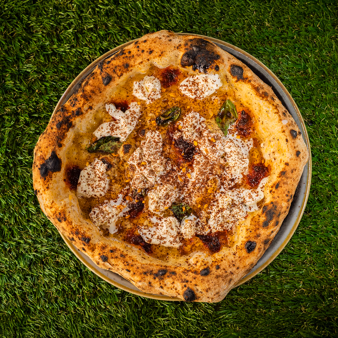 Pizza con bordi leggermente bruciati, ricoperta da fiocchi di formaggio cremoso, foglie di basilico, salsa rossa piccante e una spolverata di cacao o spezie.