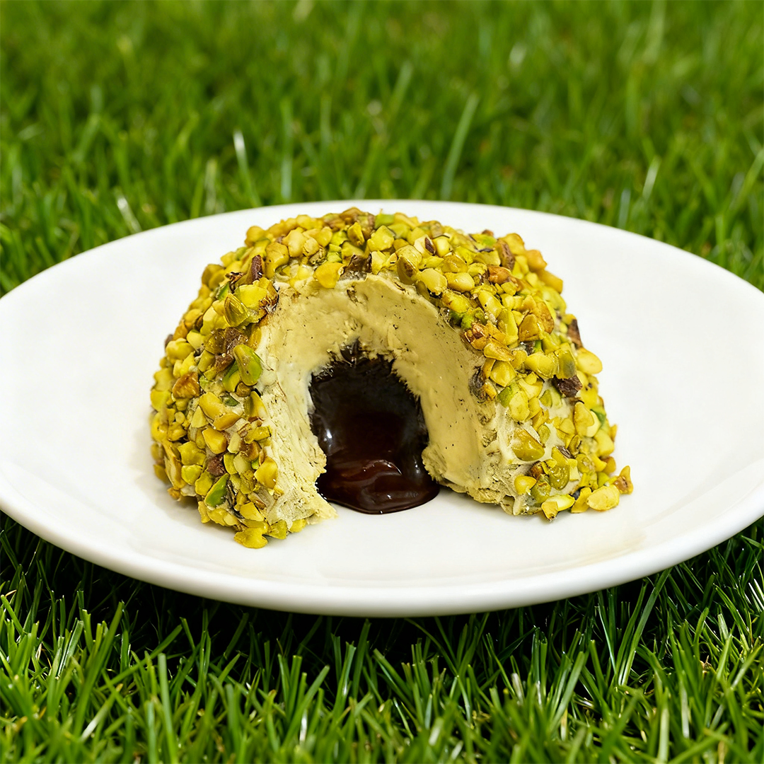 Dessert rotondo ricoperto di pistacchi tritati con un ripieno di cioccolato fuso su un piatto bianco, posato sull'erba verde.
