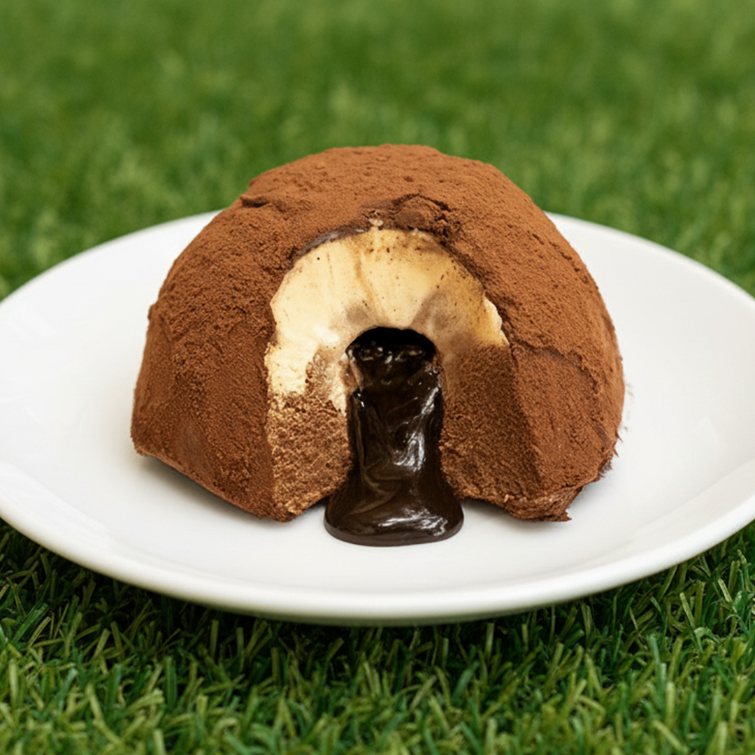 Dessert a forma di cupola con strato esterno di cacao in polvere e interno di crema chiara con cioccolato fuso che cola al centro, su piatto bianco.