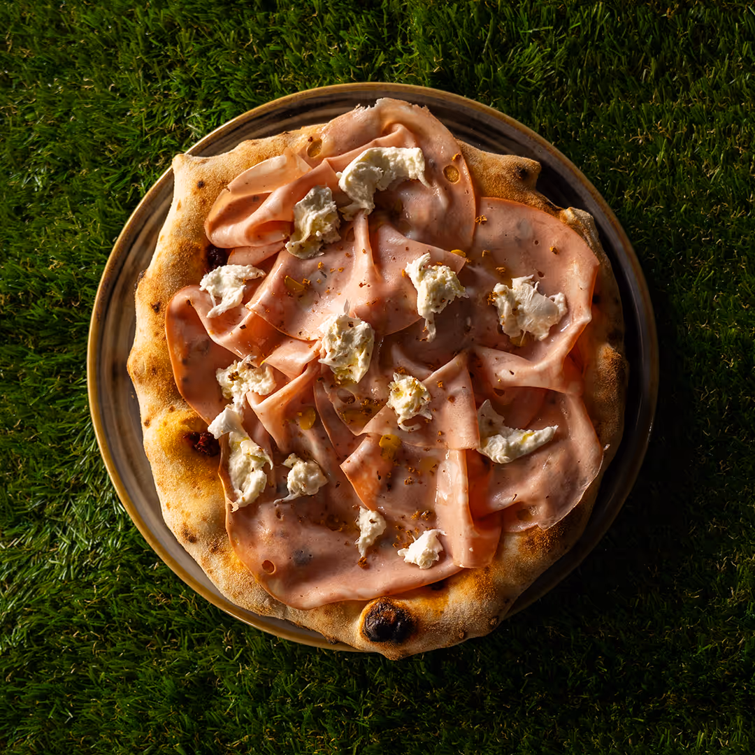 Pizza con fette sottili di mortadella e pezzetti di formaggio bianco su un piatto, su fondo di prato verde.