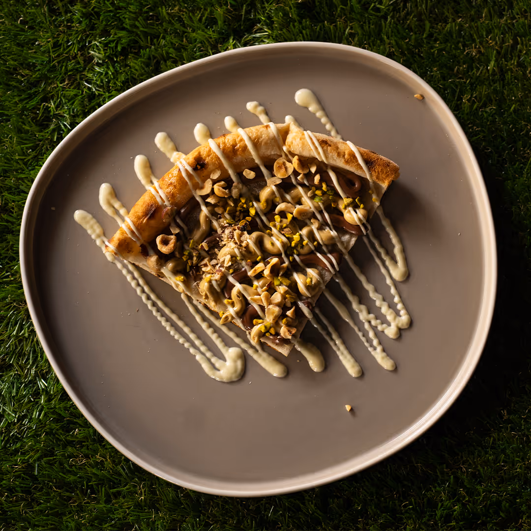Fetta di pizza dolce con crema di nocciole, granella di nocciole e pistacchi, decorata con glassa bianca su un piatto grigio su erba.