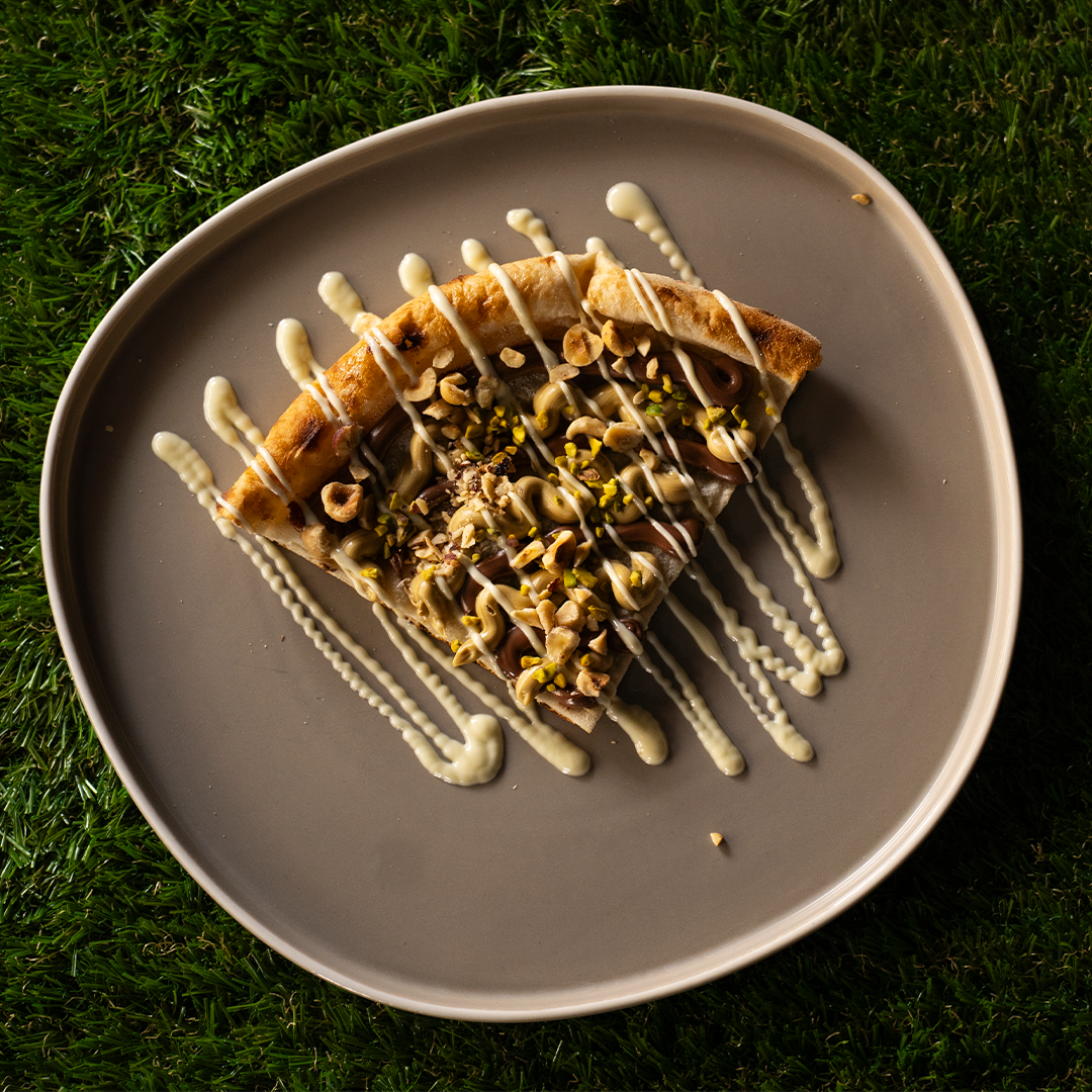Fetta di pizza dolce con crema di nocciole, granella di nocciole e pistacchi, decorata con glassa bianca su un piatto grigio su erba.