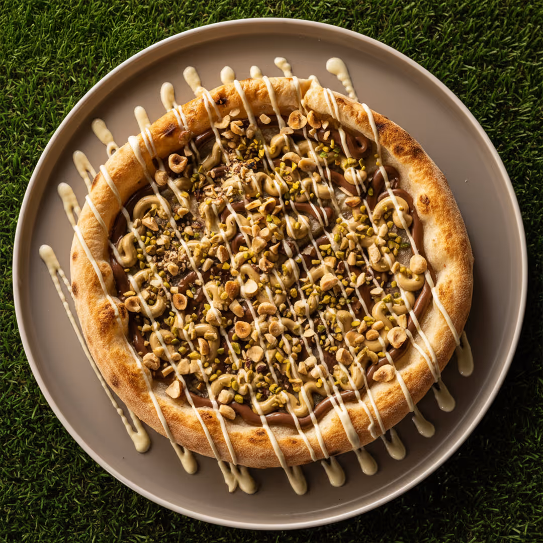 Pizza dolce con crema di cioccolato, granella di nocciole, pistacchi e glassa bianca su un piatto marrone.