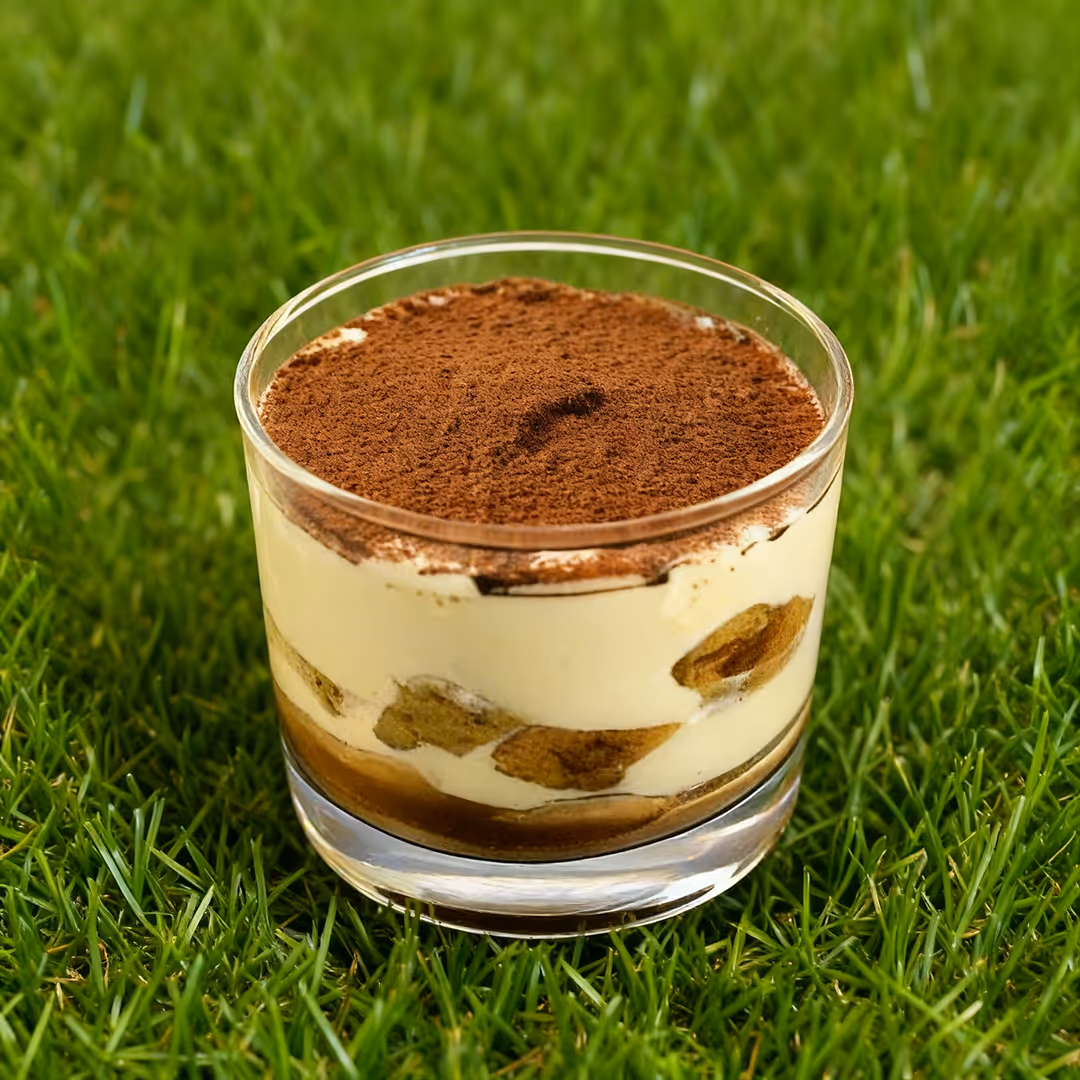 Bicchiere trasparente con tiramisĂą a strati di crema e cacao, posizionato sull'erba verde.