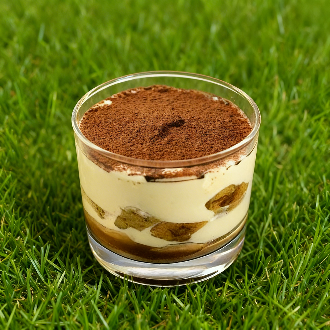 Bicchiere trasparente con tiramisù a strati di crema e cacao, posizionato sull'erba verde.