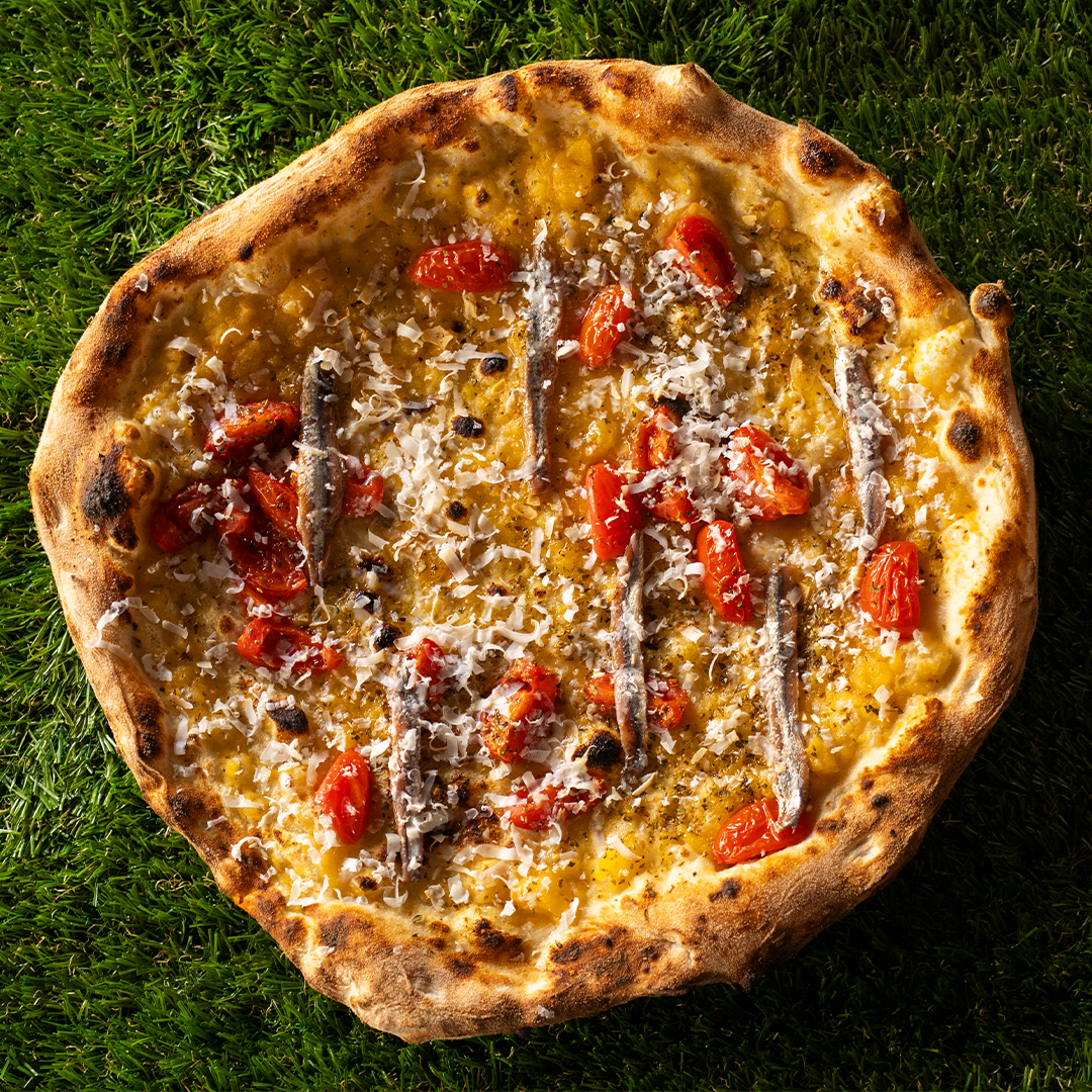 Pizza con pomodorini, acciughe, formaggio grattugiato e crosta leggermente bruciata su sfondo d'erba verde.