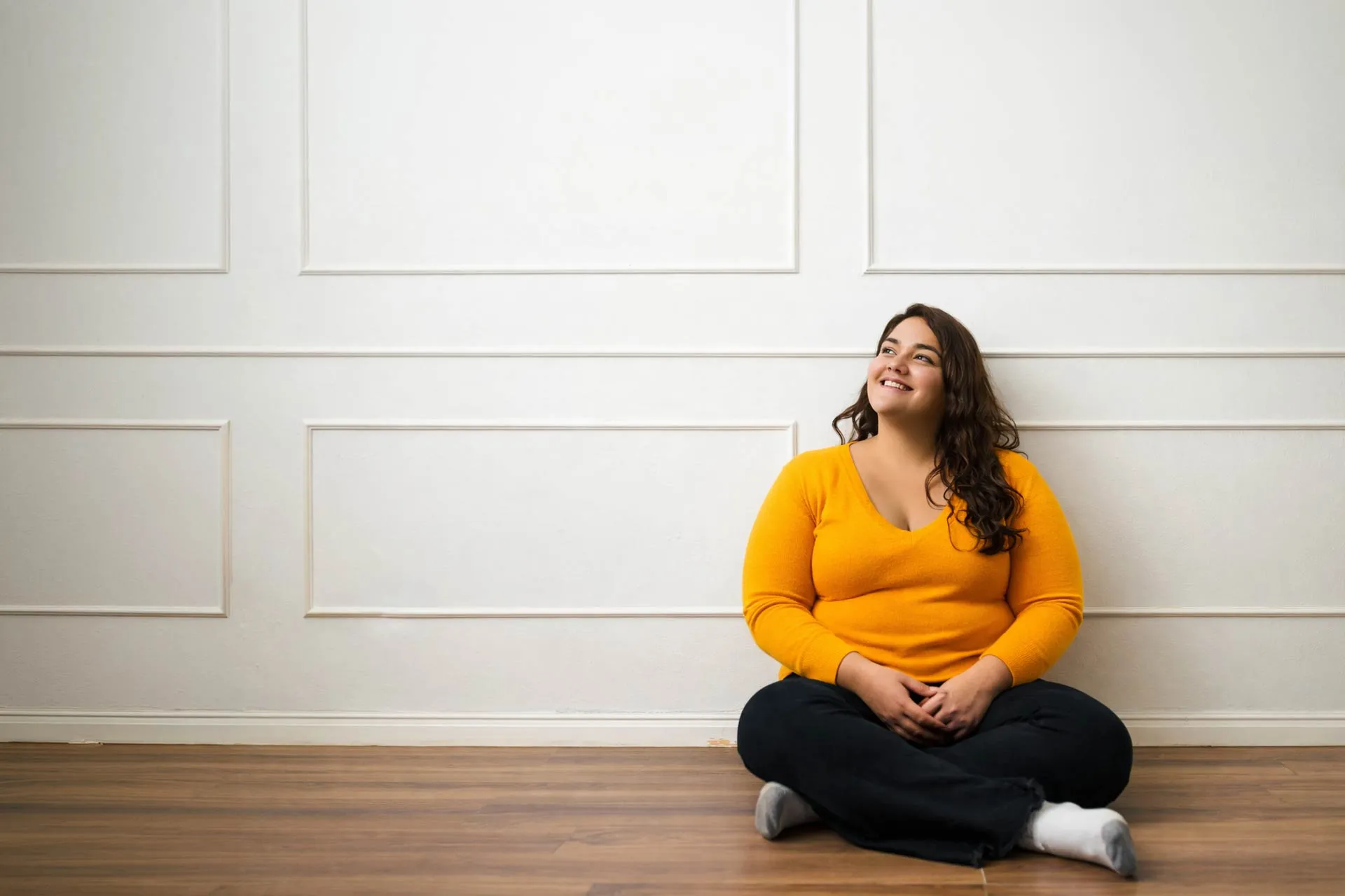Eine lächelnde Frau mit orangefarbenem Pullover sitzt mit verschränkten Beinen auf einem Holzboden vor einer weißen Wand.